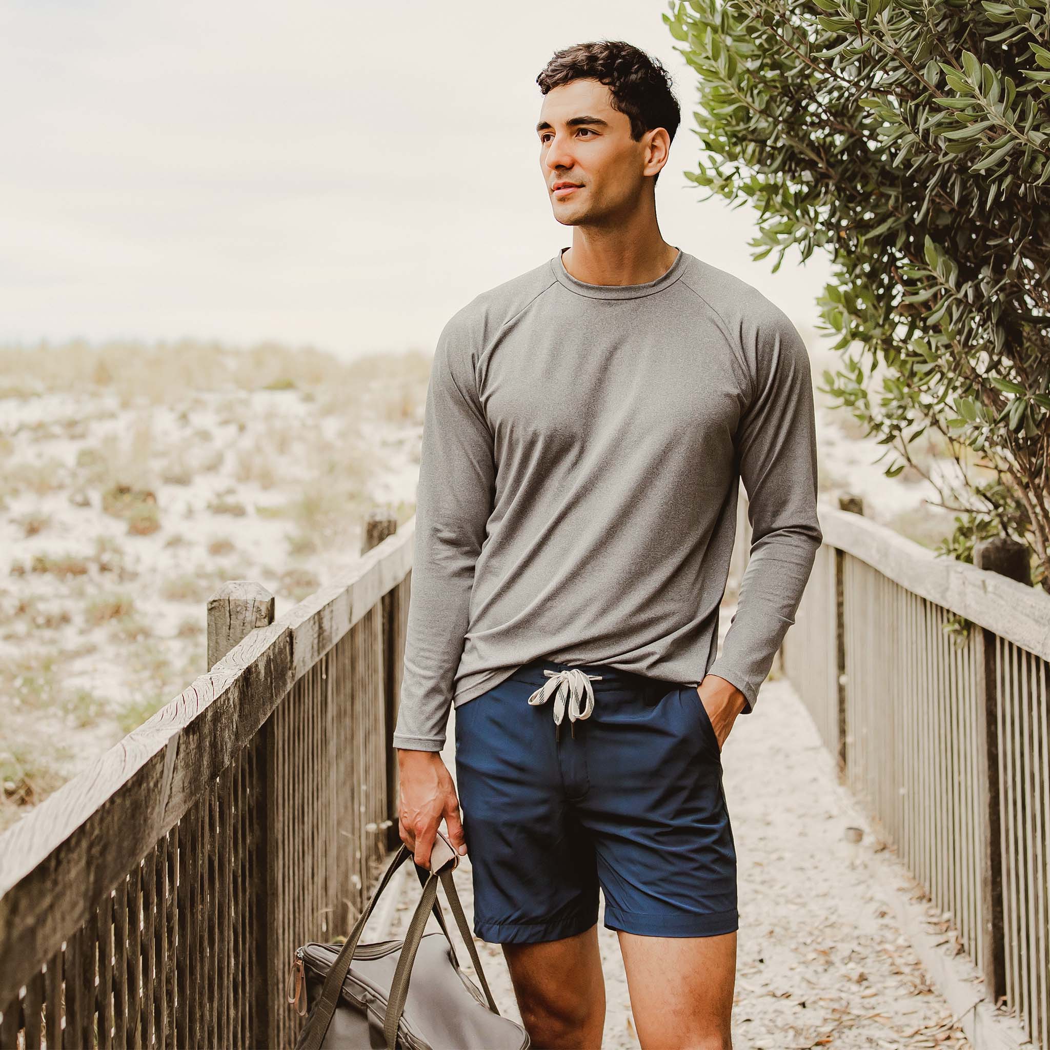 Man wearing a gray long-sleeve shirt and blue shorts standing on a wooden path with a natural background.