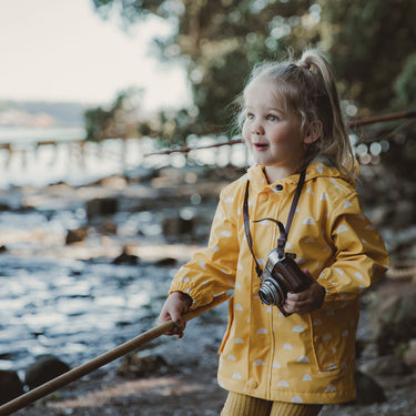 Girl holding camera and stick at beach, wearing yellow raincoat.